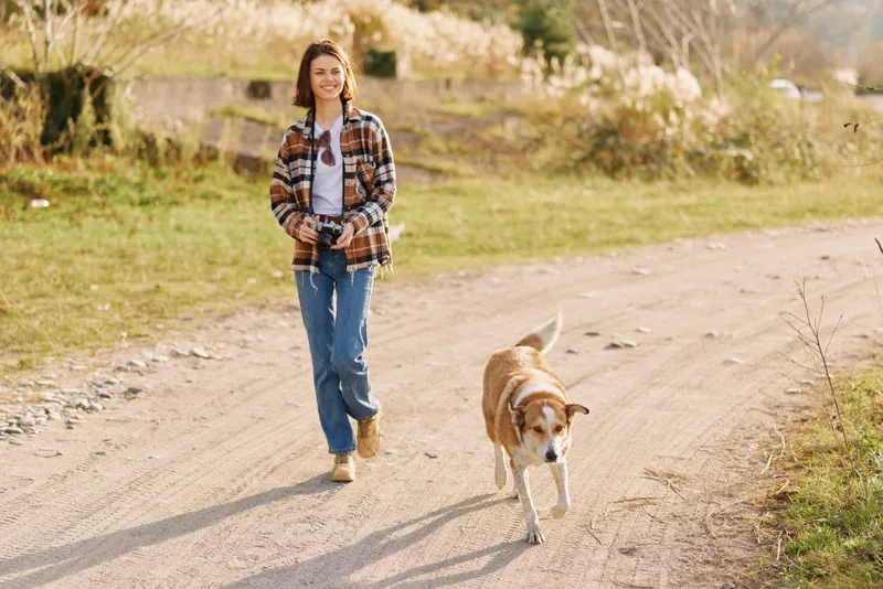 Woman walking dog outdoors on a sunny rural path, casual plaid shirt and jeans, enjoying a peaceful nature stroll and pet companionship in a relaxed daytime moment along a country trail