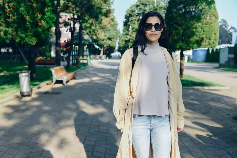 Woman wearing beige coat and gray sweater walks along a sunlit urban street, casual style and relaxed pose, fashion forward streetwear for lifestyle, travel and everyday elegance