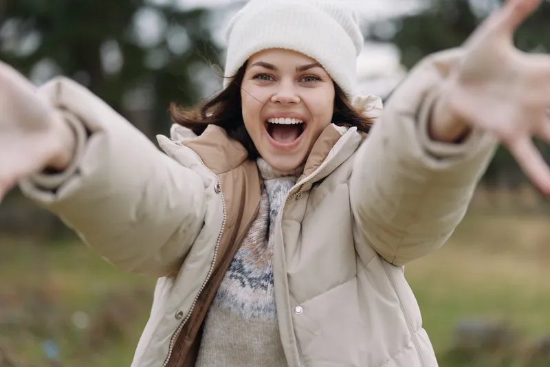 Happy woman with outstretched arms outdoors in winter, wearing a warm coat and beanie, expressing joy and invitation in a bright candid moment of spontaneous celebration