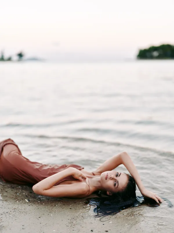 A serene portrait of a woman lying on a sandy shore beside calm water, wearing a flowing dress. soft light, gentle breeze, timeless mood, subtle elegance.