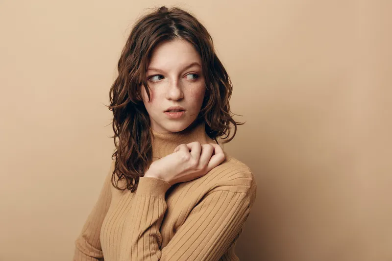 Young woman with curly brown hair wearing beige sweater looks sideways with thoughtful expression. Studio portrait on neutral background. Concept of contemplation, fashion, and natural beauty.