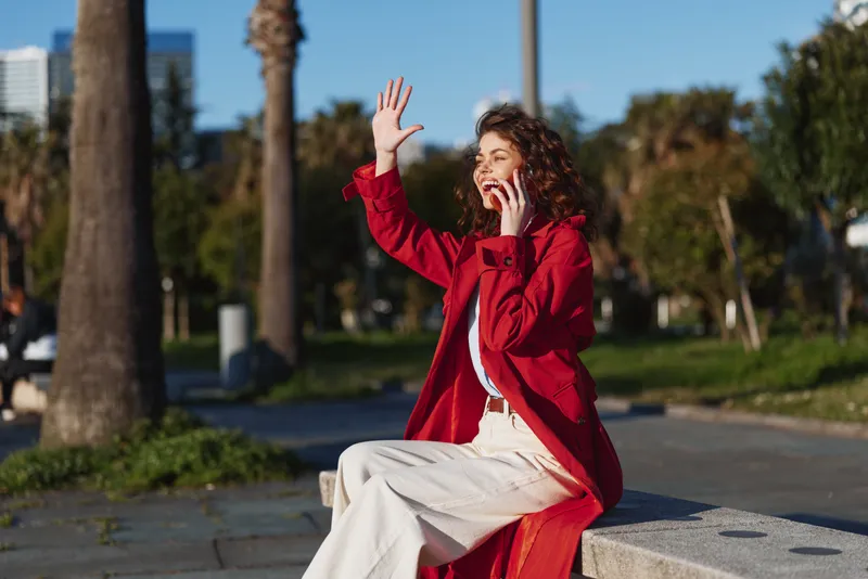 Smiling woman in a red coat sits on a bench outdoors, waving her hand. Bright daylight highlights casual fashion, joyful mood, and park scenery.