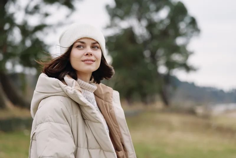 Winter woman outdoors wearing a puffy coat and beanie, standing in a calm landscape with trees, soft light, and a relaxed expression, capturing casual seasonal style