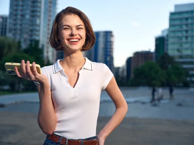 A cheerful woman in a white polo stands outdoors in an urban setting, smiling warmly while holding a smartphone, creating a bright, upbeat, professional atmosphere for a modern workspace.