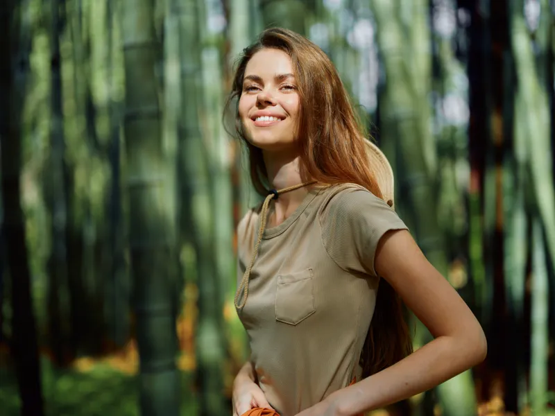 Woman portrait in bamboo forest smiling with sunlight on her face, casual outfit and backpack strap visible, nature travel scene, eco tourism, relaxed outdoor mood and warm green background