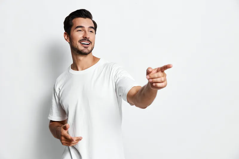 Smiling man in casual white t-shirt pointing with hand, looking at camera with cheerful expression, isolated on solid light background. Lifestyle concept.