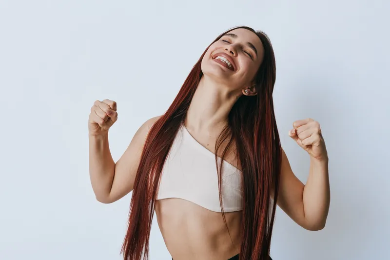 Young woman with long hair smiling and celebrating success with fists up in casual top against plain background. Emotion, happiness and confidence in studio portrait of female.