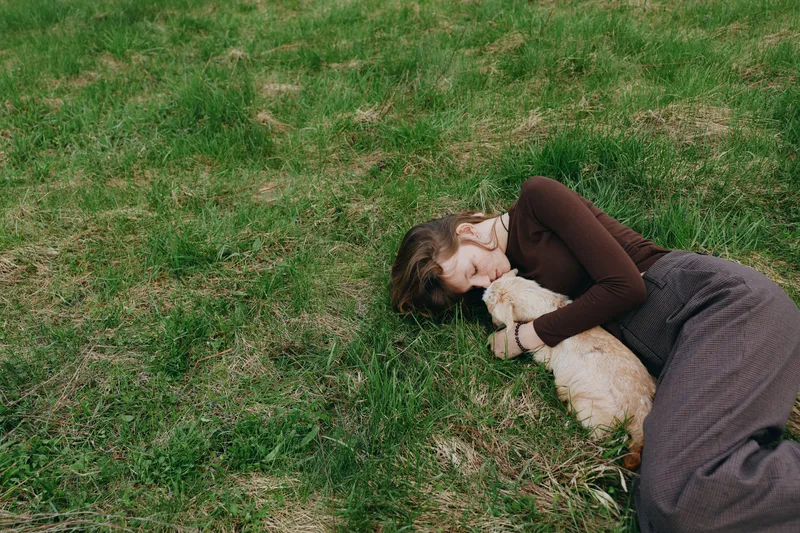 A young woman lies on green grass outdoors embracing a small baby goat while wearing a brown sweater and skirt in a peaceful lifestyle moment.