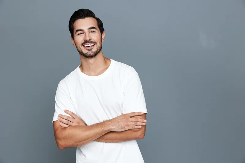 Smiling man standing with crossed arms, wearing a casual white t-shirt and looking at the camera against a plain grey background. Positive lifestyle concept.