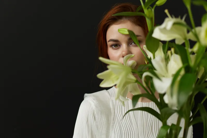 Young woman with brown hair, dressed in a white blouse, partially hidden by a bouquet of white lilies against a dark background, conveying beauty and serenity