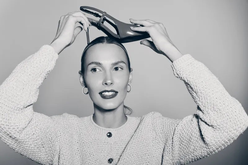 Young white woman with pale skin and dark hair holds a high heel shoe above her head, wearing a knitted sweater and hoop earrings in bright studio lighting.