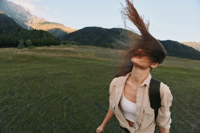 Young woman with windblown hair moves across a grassy field with distant mountains, embodying freedom, adventure, and outdoor lifestyle. Her beige shirt and white top suggest casual exploration.