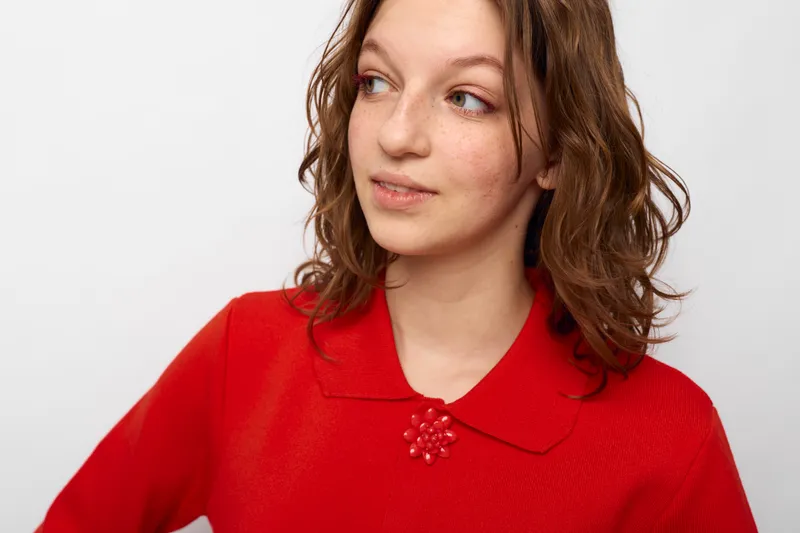Young woman with curly brown hair wearing a red sweater looks sideways with a thoughtful expression on a white background. Concept of casual style, contemplation, and natural beauty.