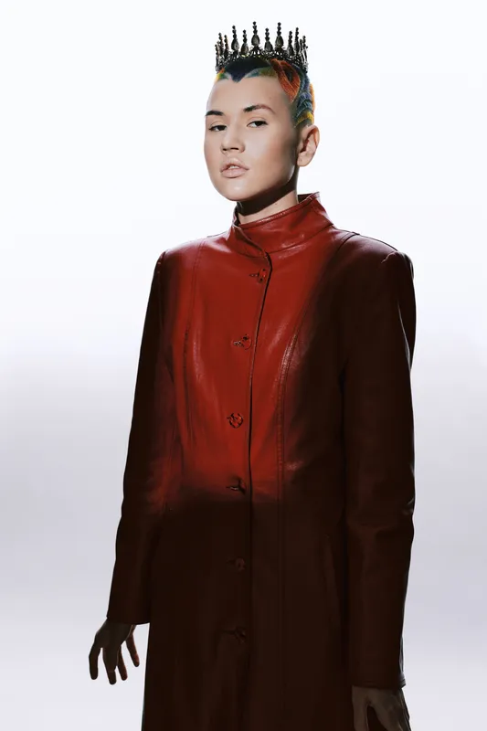 young woman with crown and colorful hair wearing red leather coat, posing confidently against white background in futuristic fashion style, portrait