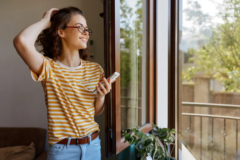Smiling woman with striped shirt standing by an open window, holding a phone. Cozy home scene highlighting casual lifestyle, natural light, and relaxed mood. Soft lighting adds warmth.
