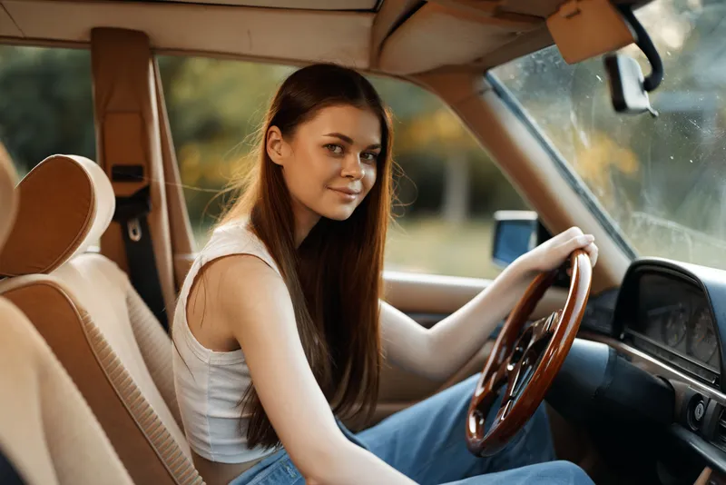 Young woman with long hair sitting in a vintage car, smiling warmly while wearing a casual white top and denim jeans, enjoying a serene drive in nature.