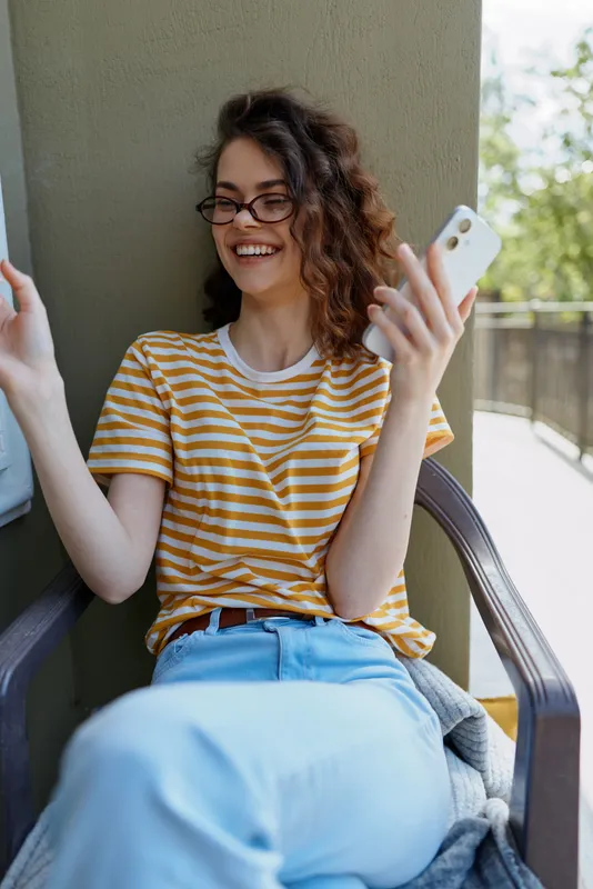 Smiling woman with striped t shirt on a balcony, holding a smartphone. Bright daylight creates a casual home mood focused on lifestyle, connection, and relaxed everyday moments.