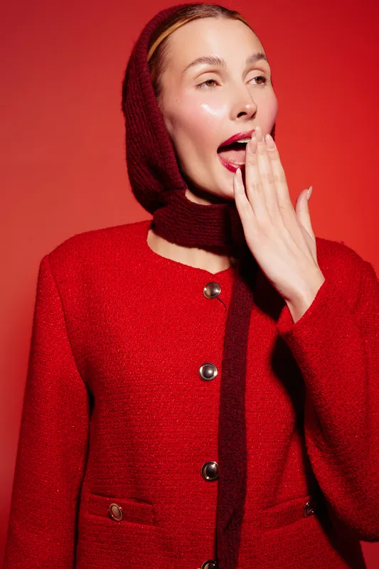 Young woman in red woolen coat and matching scarf yawns covering mouth with hand. Studio portrait on red background. Concept of tiredness, cold weather, and winter fashion.
