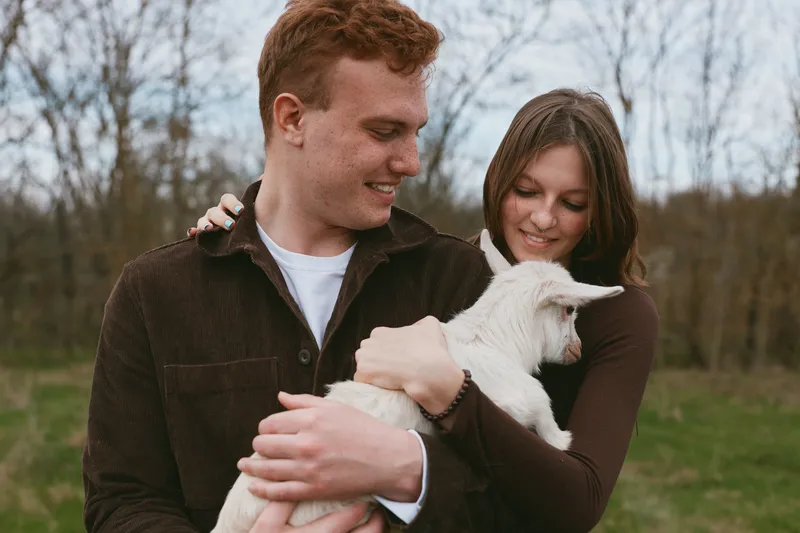 A smiling couple gently cradles a white goat kid in a grassy field, capturing a warm rural lifestyle moment of bonding, nature, and family affection, outdoors in a gentle breeze under daylight.