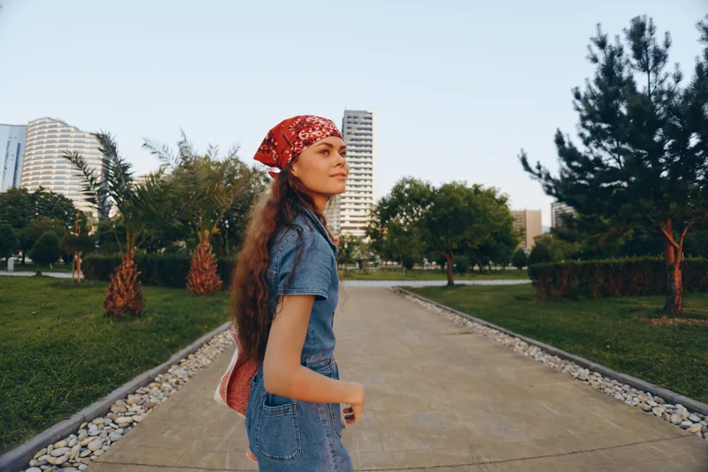 lifestyle woman in modern boho-western denim street style wearing a red bandana, walking outdoors with greenery and urban buildings in film color tones