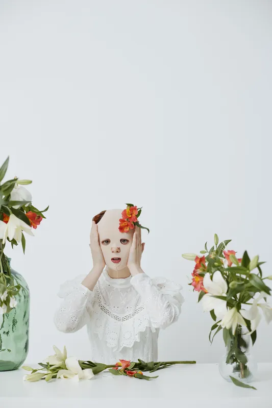 A young woman in a white dress with floral accents looks distressed while holding her head, surrounded by flowers, on a minimalist white background
