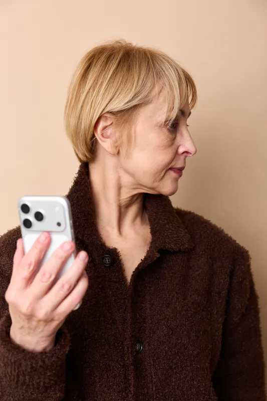 Profile portrait of elderly woman holding smartphone for communication on beige background, focused expression, technology concept, soft studio light, calm mood