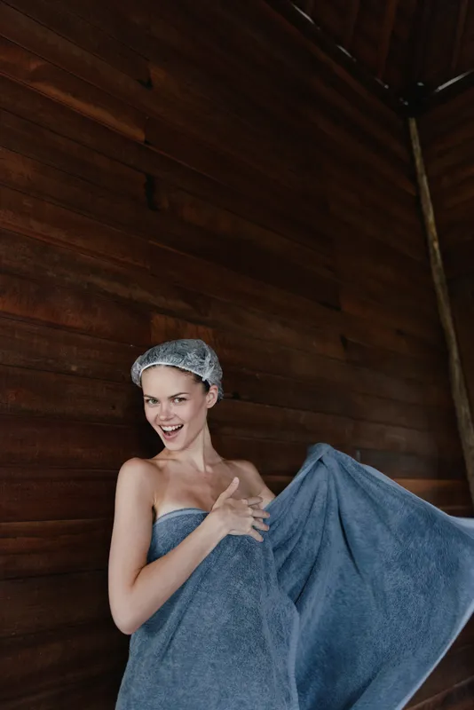 Smiling woman wrapped in blue towel with shower cap stands against wooden wall, showing joyful and playful expression indoors in warm spa environment.