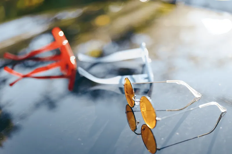 A close view shows sunglasses resting on a reflective surface with a bright outdoor scene blurred in the background, creating a casual lifestyle moment and a warm, sunny mood.