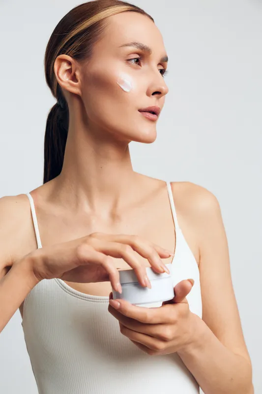 A young caucasian woman calmly applies face cream to her cheek in a clean studio, demonstrating a focused skincare routine, with a jar of moisturizer in her hand and natural lighting.