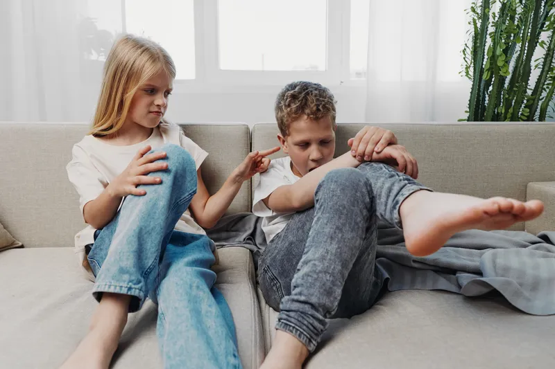 Two children sitting on a sofa in a bright living room, a girl pointing her finger at a boy who looks upset, capturing sibling interaction and emotional conflict at home.