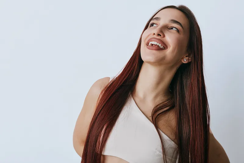 Young woman with long straight hair smiling and looking up on white background. Portrait of happy female with natural makeup wearing sleeveless top and earrings.