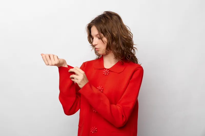 Young woman in red sweater adjusts sleeve while looking down. Casual style, indoor studio with white background. Concept of fashion, comfort, and everyday clothing.