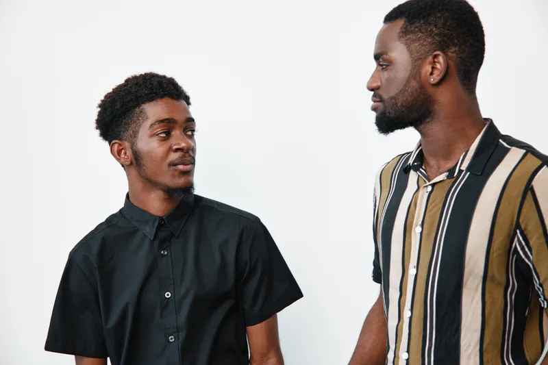 Two african american men with dark skin standing and looking at each other in casual shirts against white background. Young black males with natural hair and facial hair indoors.