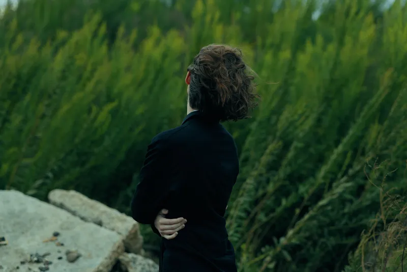 Businessman with back turned in a dark suit outdoors by a rocky shore, contemplative silhouette amid grasses, emphasizing reflection, calm mood and growth