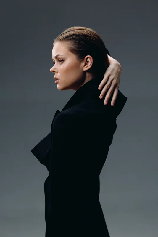 Profile of serious woman with slicked-back hair wearing black coat against dark background in studio