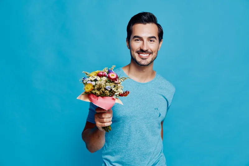 Smiling happy young man holding bouquet of colorful flowers isolated on bright blue background. Joyful celebration, love, gratitude, or special occasion concept.