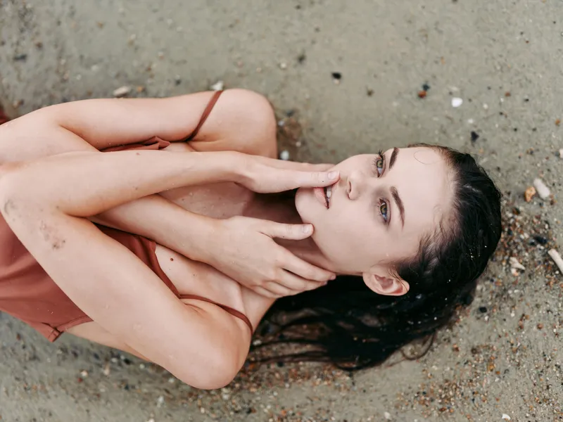 A woman lies on a sandy beach, hands gently cradling her face, conveying vulnerability, introspection, and quiet emotion in a soft, sunlit coastal setting.