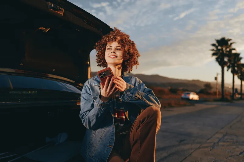 Woman with curly hair wearing denim jacket, sitting beside a parked car at sunset, smiling with hands near her face, outdoor roadside lifestyle, warm golden light, relaxed evening travel mood