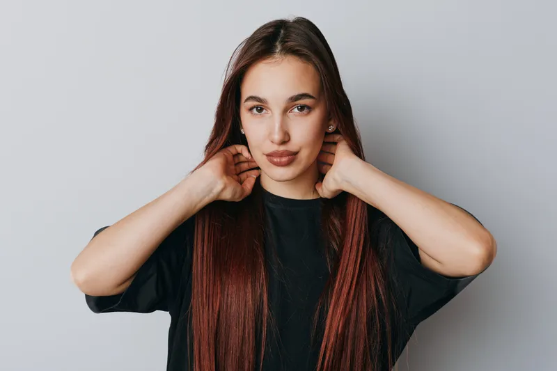 Young woman with long straight hair posing with hands near neck wearing black shirt on plain background. Casual style, confident expression and natural makeup for portrait photography.