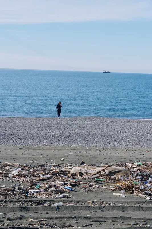 Beach scene with a lone person standing near the water, foreground litter and debris on the shore, calm sea and distant boat on the horizon, clear blue sky above.