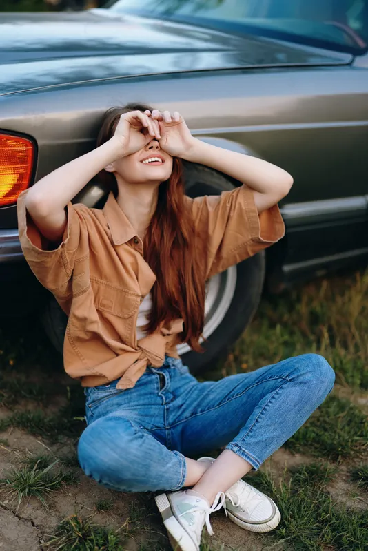 Young woman laughing with closed eyes, sitting on the grass beside a vintage car, wearing a casual oversized shirt and jeans, showcasing joy and carefree summer vibes.