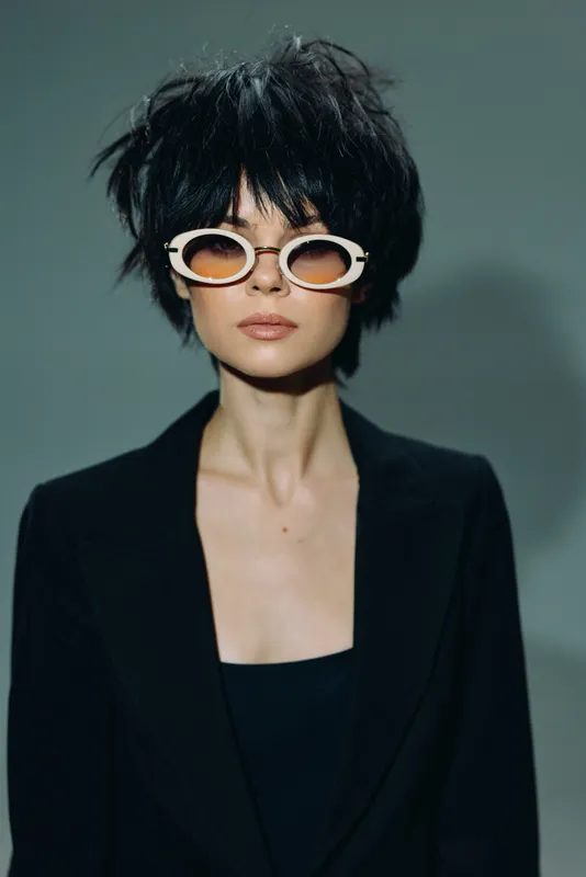 Stylish woman with short black hair wearing white sunglasses and a black blazer, posing confidently in a minimalistic setting.