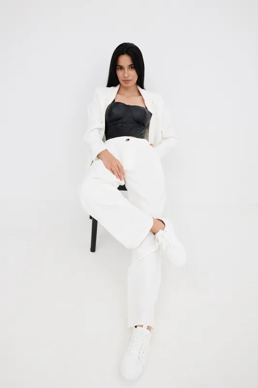 Young woman sitting confidently dressed in white pants and jacket paired with a black corset top and white sneakers against a plain light background.