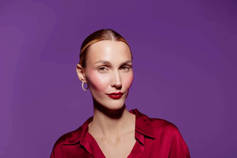 Portrait of confident woman with slicked hair wearing red blouse and gold hoop earrings on purple background. Concept of fashion, beauty, elegance, and modern style.