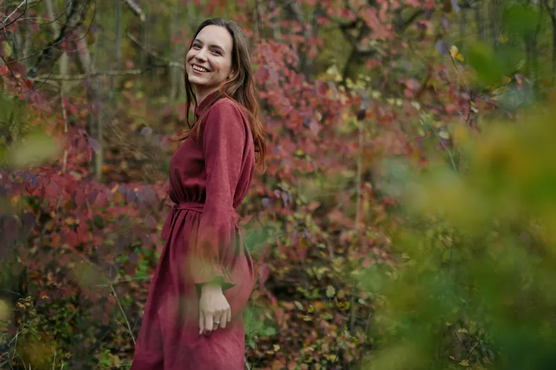 A candid portrait of a woman in a burgundy dress in autumn woodland, conveying authenticity and credibility through natural light and a relaxed, genuine pose.