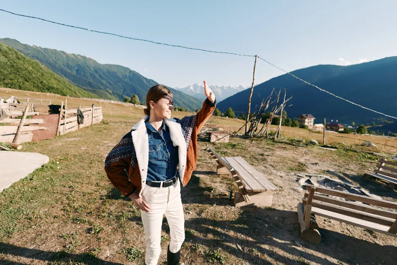 Woman in mountains rural outdoors waving in meadow, casual shearling jacket and denim shirt, standing on country path by wooden fences and benches under bright sunny alpine valley sky
