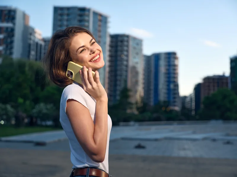 A cheerful woman stands outdoors in an urban setting, talking on a mobile phone. She smiles brightly, enjoying a casual moment amid modern buildings and clear skies.