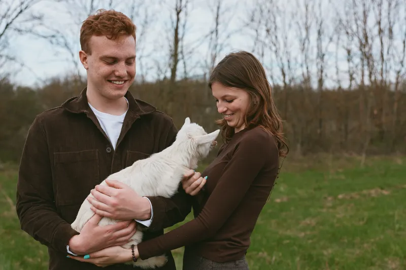 A happy couple outdoors holds a white goat kid, sharing a tender moment. A lifestyle scene focusing on family bonding with animals in a rural setting.