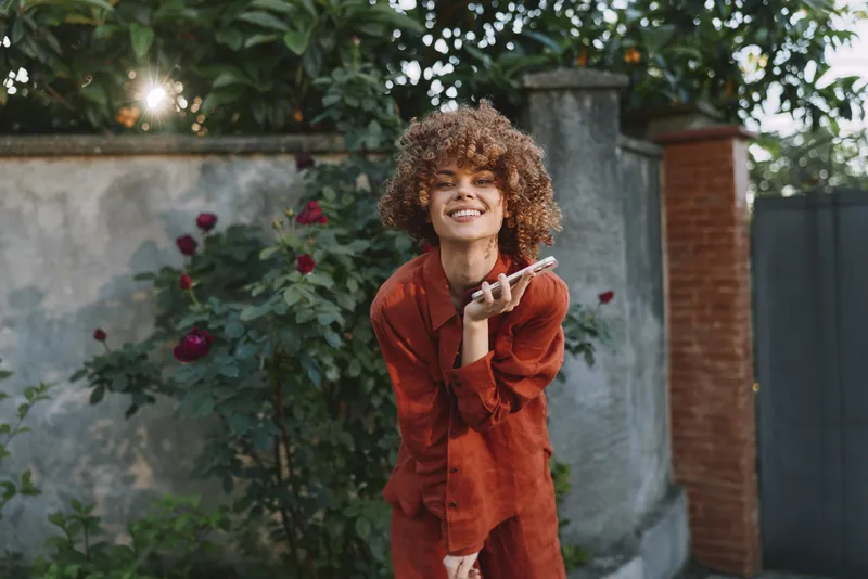 Smiling woman with curly hair wearing a red dress outdoors in a garden setting, blowing a kiss toward the camera, radiating joyful lifestyle, natural light, vibrant mood, and relaxed confidence