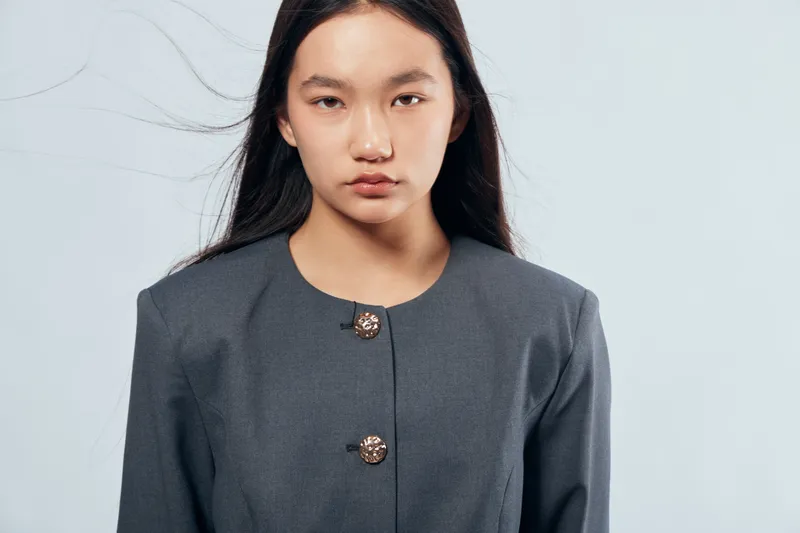 Young woman with long dark hair wearing a gray jacket with decorative buttons against light background looking seriously at camera, studio portrait with natural expression.